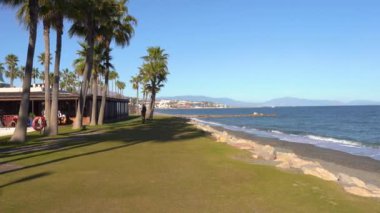 Marbella, İspanya - 24 Ocak 2024 - Palmiye ağaçları, çimenli bir çim, deniz ve dağ manzaralı sahil geçidi.