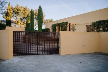 Kırpılmış kapı, sarı duvarlar, garaj yolu, selvi ağaçları, çiçek açan sarmaşıklar, berrak gökyüzü..