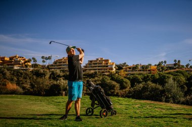 Sotogrante, İspanya - 25 Ocak 2024 - Golfte sallanan adam, havada top, yanında itme arabası, arka planda binalar, açık gökyüzü.