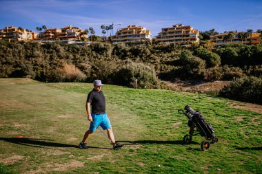 Sotogrante, İspanya - 25 Ocak 2024 - Golf arabasıyla golf sahasında yürüyen bir golfçü, arka planda konut binaları, açık gökyüzü.