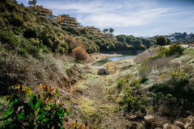 Sotogrante, İspanya - 25 Ocak 2024 - Çevresi bitki örtüsüyle çevrili küçük bir göl, açık bir gökyüzünün altındaki tepenin üzerinde yerleşim binaları.