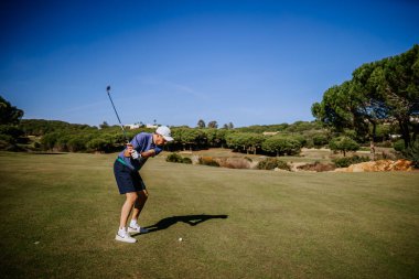 Sotogrante, İspanya - 25 Ocak 2024 - Mavi kıyafetli bir golfçü, arka planda açık mavi gökyüzü ve ağaçlı bir çim sahasında golf sopası sallıyor.
