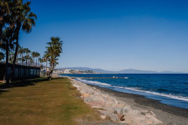 Sotogrante, İspanya - 25 Ocak 2024 - Çimenli kenar boyunca uzanan palmiye ağaçları, çakıl taşı plajı, mavi deniz ve berrak gökyüzünün altındaki uzak dağlar.