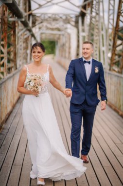 Valmiera, Letonya - 7 Temmuz 2023 - Bir gelin ve damat eski bir köprüde el ele yürüyorlar, gülümsüyorlar, arka planda yeşillik var.