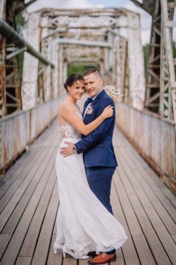 Valmiera, Letonya - 7 Temmuz 2023 - Bir gelin ve damat kırsal bir köprüye sarılmış, gülümsüyor ve elinde buketle kameraya bakıyor.