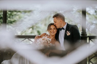 Valmiera, Letonya - 7 Temmuz 2023 - Damat gelini yanaktan öpüyor, hem gülümsüyor, hem çardakta oturuyor, bulanık beyaz ön plan, romantik ortam