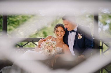 Valmiera, Letonya - 7 Temmuz 2023 - Gülen gelin oturdu, damat yakın durdu, hem düğün kıyafeti içinde, hem ön planda bulanıklık, açık hava çardağı