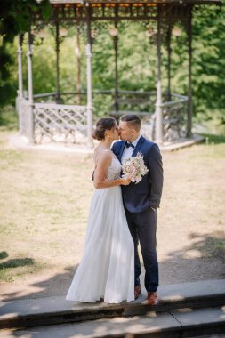 Valmiera, Letonya - 7 Temmuz 2023 - Gelin ve damat, arka planda bir çardak ve ağaçlarla kucaklaşıp öpüşüyor