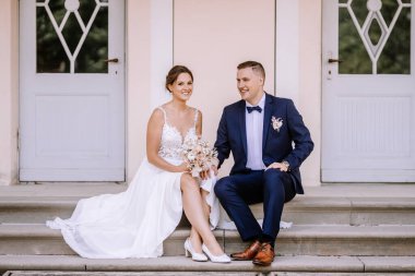 Valmiera, Letonya - 7 Temmuz 2023 - Gelin ve damat merdivenlerde oturuyor, birbirlerine gülümsüyorlar, arkalarında geometrik pencereler olan bir kapı.