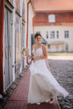 Valmiera, Letonya - 7 Temmuz 2023 - Çiçekli, akıcı beyaz elbiseli gelin, kaldırım taşı bir sokakta yürüyor, arka planda binalar var.