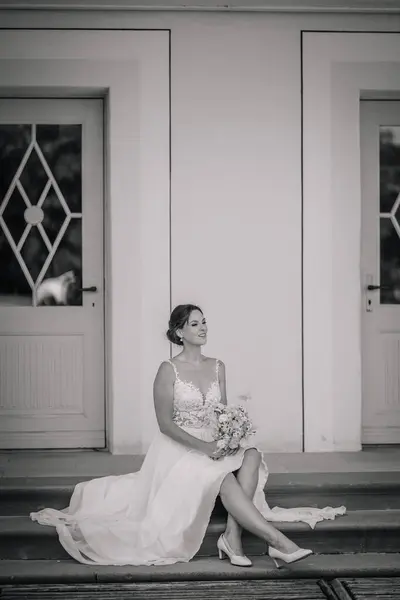 Valmiera, Latvia - July 7, 2023 - Bride seated on steps in monochrome, looking to the side, with double doors in the background.