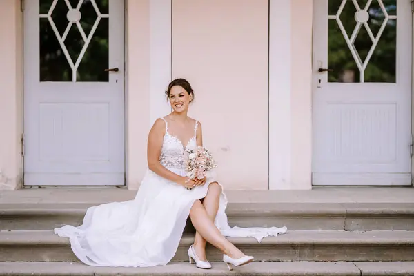 Valmiera, Latvia - July 7, 2023 - Bride seated on steps in front of double doors, smiling at camera, holding a bouquet.