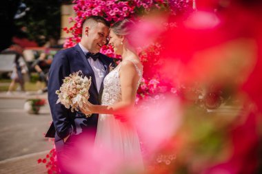 Valmiera, Letonya - 7 Temmuz 2023 - Bir gelin ve damat birbirlerine gülümsüyor, ön planda parlak pembe bir çiçekle çerçevelenmiş.