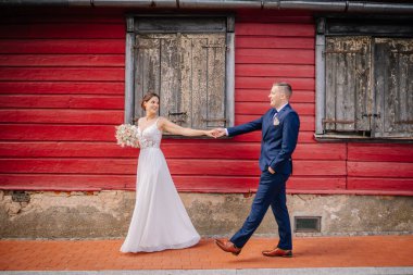 Valmiera, Letonya - 7 Temmuz 2023 Gelin ve damat kırmızı ahşap bir duvarın önünde el ele tutuşmuş, gelin gülümsüyor ve geriye bakıyor.