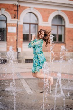 Valmiera, Letonya - 7 Temmuz 2023 - Mavi-yeşil elbiseli neşeli bir kadın, fıskiyelerden gelen su jetlerinin arasında, arka planda tarihi bir tuğla bina ile yakalandı.