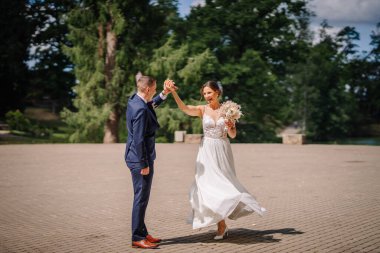 Valmiera, Letonya - 7 Temmuz 2023 - Güneşli bir günde gelin ve damat dans ediyor, damat mavi takım elbise içinde, gelin elinde buket, arka planda ağaçlar