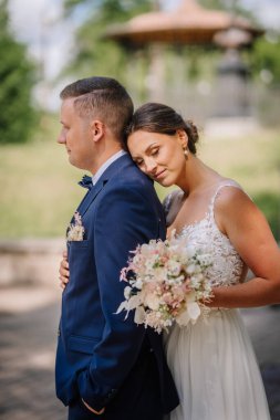 Valmiera, Letonya - 7 Temmuz 2023 - Gelin damadın omzuna yaslanıyor, ikisi de düğün kıyafeti giymiş, elinde buket, açık havada, arka planda çardak, samimi, barışçıl, sevecen.