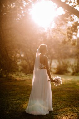 Valmiera, Letonya - 7 Temmuz 2023 - Beyaz elbiseli bir gelin, elinde bir buket, ağaçların arasından güneş tarafından aydınlatılan bir buketle uzaklara bakar..