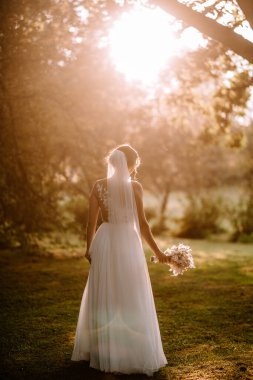 Valmiera, Letonya - 7 Temmuz 2023 - Arkadan bir gelin, elinde bir buket, ağaçların arasından süzülen güneş ışığıyla.