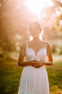 Valmiera, Letonya - 7 Temmuz 2023 - Beyaz dantelli bir gelinlik giymiş ışıldayan bir gelin güneşli bir parkta ellerini sıkarak duruyor.
