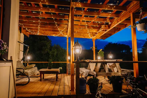 Valmiera, Riga - 5 Temmuz 2023 - Alacakaranlıkta sicim ışıklarıyla süslenmiş, rahat koltuklar ve saksı bitkileriyle dolu açık hava ahşap bir güverte.