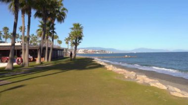 Sotogrande, İspanya - 23 Ocak 2024 - Palmiye ağaçları, çimenler, sahil, deniz ve bir binayla kıyı manzarası.