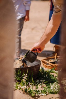 Jurmala, Letonya - 25 Temmuz 2023 - Bir kişi siyah bir bardaktan sıvı döker yeşil ve beyaz çiçeklerden oluşan bir yatağa, halk unsurlarının görüldüğü bir plaj ayininin bir parçası olan.