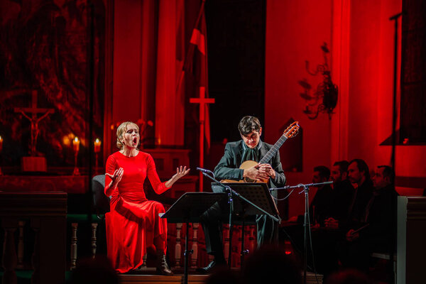 Valmiera, Latvia - January 7, 2024 - Saint Simon's Church. Soprano in red singing with classical guitarist performing in church, red lit background, audience in foreground.