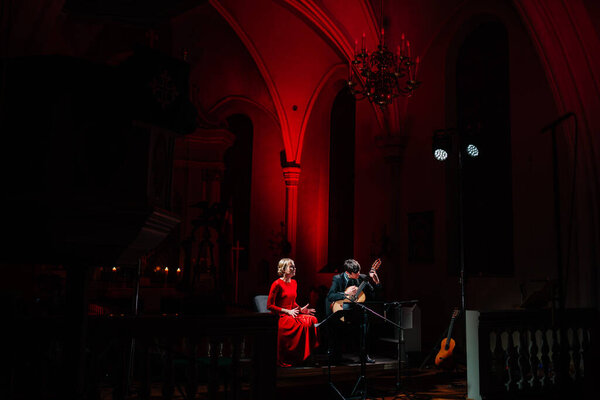 Valmiera, Latvia - January 7, 2024 - Saint Simon's Church. Musicians on stage in a church, woman in red dress and man with guitar, red lighting on arches, chandelier above.