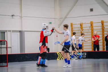 Valmiera, Letonya - 17 Şubat 2024 - Sahada iki florbol oyuncusu, biri kaleci kıyafeti giymiş, diğer oyuncularla birlikte arka planda bir antrenör var..