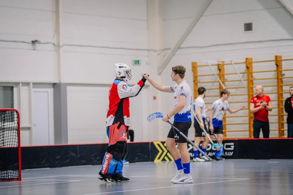 Valmiera, Letonya - 17 Şubat 2024 - Sahada iki florbol oyuncusu, biri kaleci kıyafeti giymiş, diğer oyuncularla birlikte arka planda bir antrenör var..