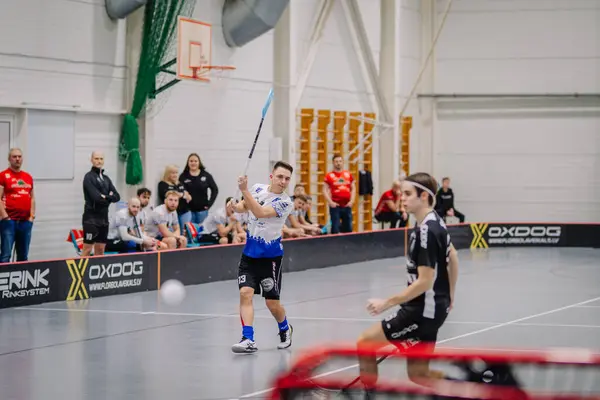 Valmiera, Letonya - 17 Şubat 2024 - Beyaz formalı bir florbol oyuncusu maç sırasında sopasıyla yüksek vuruş yapıyor, arka planda seyirciler var..