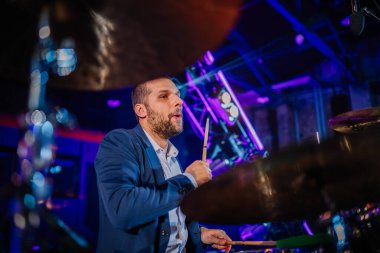 Riga, Letonya - 16 Şubat 2024 - Konserde çalan odaklanmış bir davulcu, sahne ışıkları ve zilleri görünür.