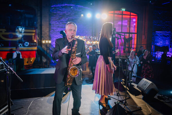 Riga, Latvia - February 16, 2024 - A saxophonist playing alongside a female singer on stage at a vibrant event with guests and colorful lighting in the background.