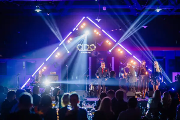 Riga, Letonya - 16 Şubat 2024 - Bir şarkıcı, bir klavyeci ve vokalist de dahil olmak üzere canlı bir mavi sahne ışıklandırmasıyla sahnede bir grup sahne alır..