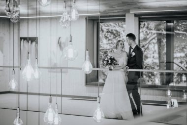 Riga, Letonya - 20 Ocak 2024 - Bir gelin ve damat birbirlerine gülümsüyor, orman manzaralı bir odada asılı ampullerle çevrili..