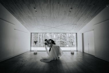 Riga, Letonya - 20 Ocak 2024 - Gelinin elbisesi akarken, karlı bir orman zeminli minimalist bir odada öpüşen bir çift.