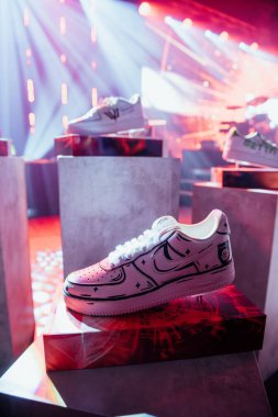 Riga, Latvia - December 18, 2023 - pair of white sneakers with black detailing displayed on a pedestal with a vibrant red and white light show in the background. clipart