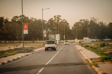 Agadir, Fas - 28 Şubat 2024 - araçlar, alacakaranlıkta ağaçlarla kaplı bir caddede, kaldırımda, yumuşak altın bir gökyüzünün altında, bir yayalar ve reklam pankartlarıyla dolaşırlar.