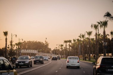Agadir, Fas - 28 Şubat 2024 - Gün batımında palmiye çizgili bir bulvarda trafik akıyor, sokak lambaları ve arka planda puslu bir gökyüzünün altında yaya köprüsü var..
