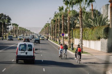 Agadir, Fas - 28 Şubat 2024 - Bisikletçiler ve araçlar ağaçlık bir yolu paylaşıyor; trafik ışıkları, sokak lambaları ve manzaralı bir kaldırım görünür.