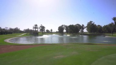 Agadir, Fas - 27 Şubat 2024 - Büyük bir su tehlikesi, fıskiyeler, kum sığınakları, palmiye ağaçları ve açık mavi gökyüzü ile panoramik bir golf sahası manzarası.