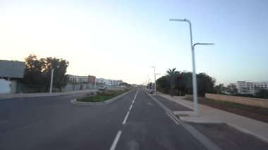 Agadir, Fas - 25 Şubat 2024 - Bir otobüsten yol manzarası, hava kararırken ya da şafak sökerken gökyüzünün altında binaların ve palmiye ağaçlarının olduğu boş bir caddeyi gösteriyor.