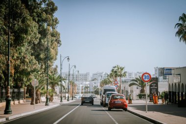 Agadir, Fas - 29 Şubat 2024 - Ağaçlarla çevrili bir şehir bulvarı içinde arabalar, palmiye ağaçları, sokak lambaları, hız limiti işaretleri ve açık mavi gökyüzü, uzaktaki beyaz binalara doğru gidiyor..