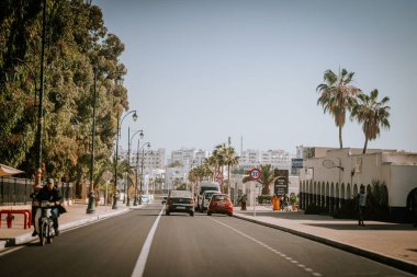 Agadir, Fas - 29 Şubat 2024 - Palmiye ağaçları, sokak lambaları, motosikletçiler, arabalar ve binalarla kaplı güneşli bir şehir caddesi, hız limiti işaretleri ve açık gökyüzü