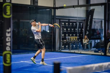 Riga, Letonya - 3 Mart 2024 - Şapkalı bir erkek oyuncu kapalı sahada bir padel topuna vurmak üzere, kupalar ve bulanık arka planda görünen bir izleyici.