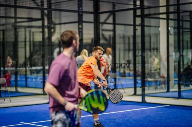 Riga, Letonya - 3 Mart 2024 - Kapalı bir tesiste iki adam padel oynuyor; turuncu gömlekli biri raketiyle topa vurmaya hazır.
