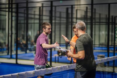 Riga, Letonya - 3 Mart 2024 - Padel mahkeme ağında üç adam sohbet ediyor, ikisi raket tutuyor, biri bir şey açıklıyormuş gibi hareket ediyor, kapalı bir alanda.
