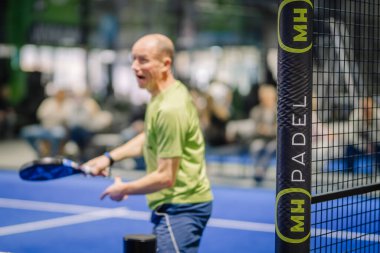Riga, Letonya - 3 Mart 2024 - Yaşlı bir adam aktif olarak padel oynuyor, iç mekanda bulanık bir seyirciyle yüz ifadeleriyle oyun oynarken yakalandı..