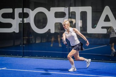 Riga, Letonya - 3 Mart 2024 - Spor kıyafetleri içinde odaklanmış bir kadın mavi bir salonda padel oynuyor, arkasında büyük beyaz harfler var..